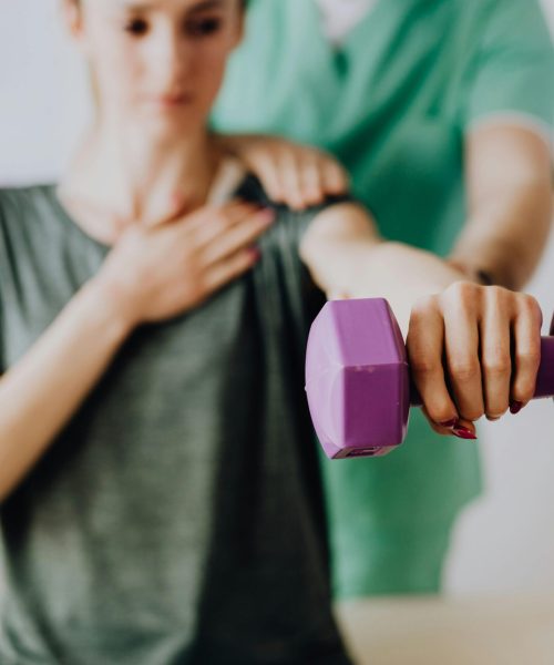 Crop unrecognizable orthopedist in uniform helping slender female in casual wear reaching arm with dumbbell in doctor office in hospital